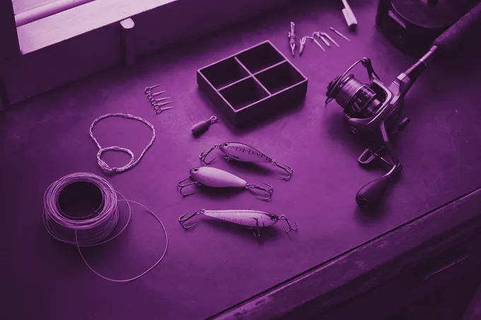Fishing tackle on a dark workbench in grape duotone: three crankbait lures with treble hooks, a baitcasting reel, a spool of line and an open compartment tray.