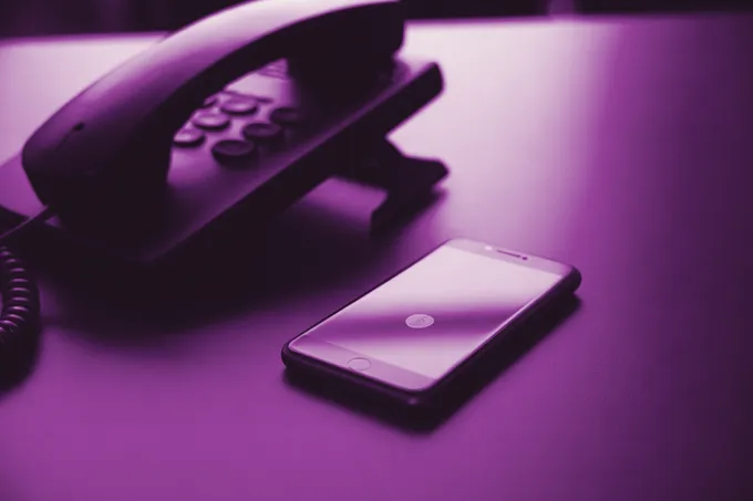 A smartphone face up on a desk beside a telephone handset off its cradle, the screen glowing, in grape duotone.